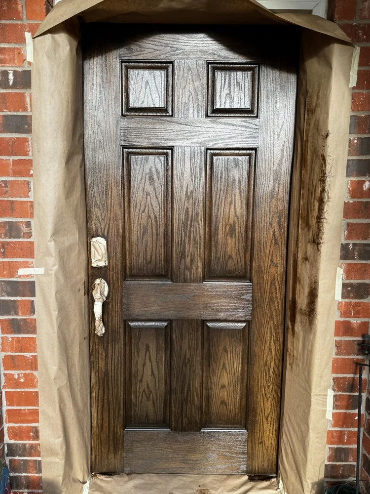 Six-panel front door freshly stained a rich dark wood tone, surroundings masked off